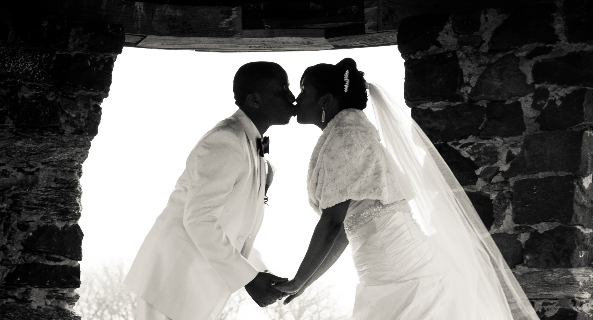 A bride and groom kiss in silhouette beneath a stone arch