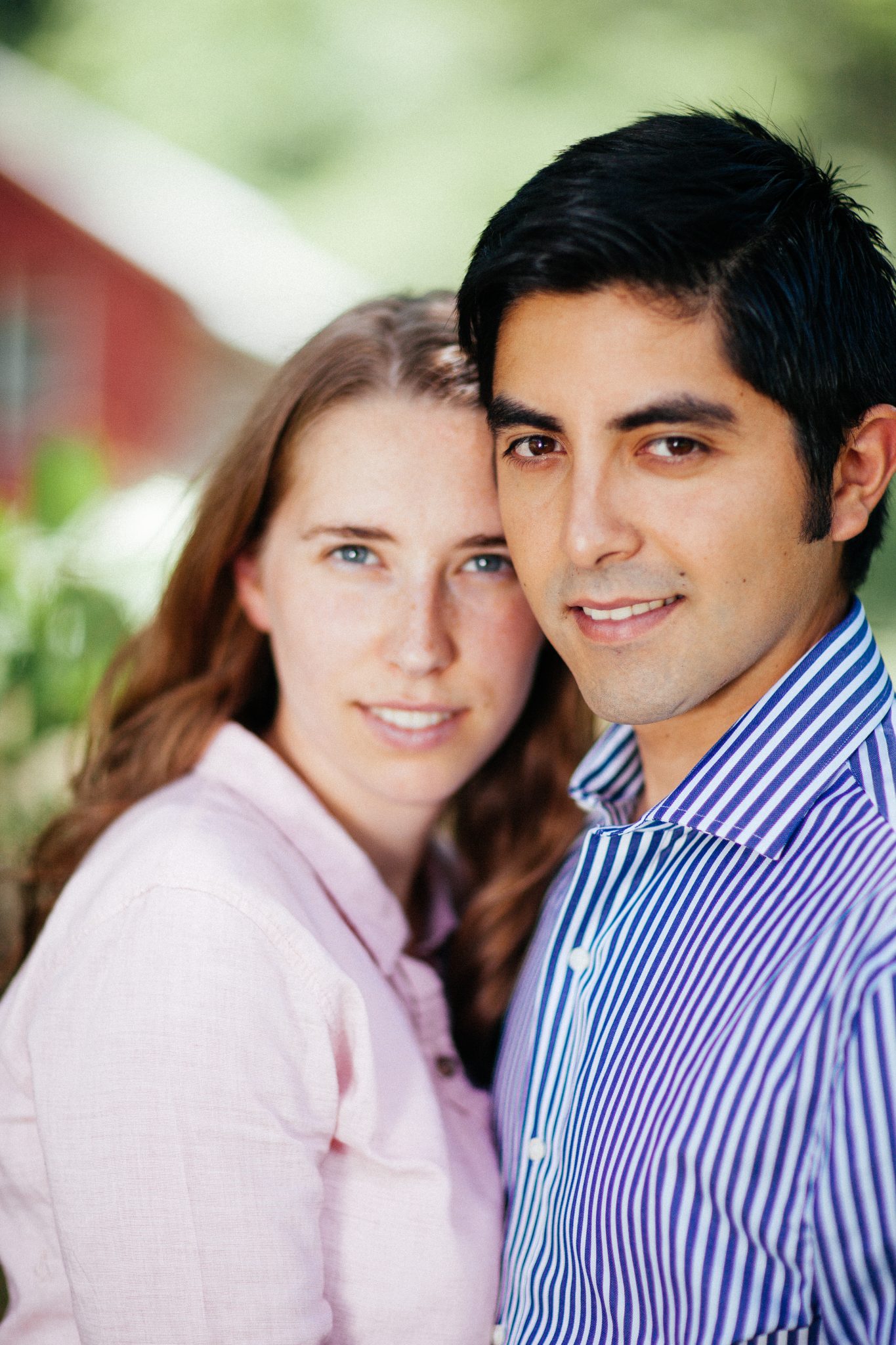 A couple cheek to cheek in soft daylight, a red barn blurred behind them