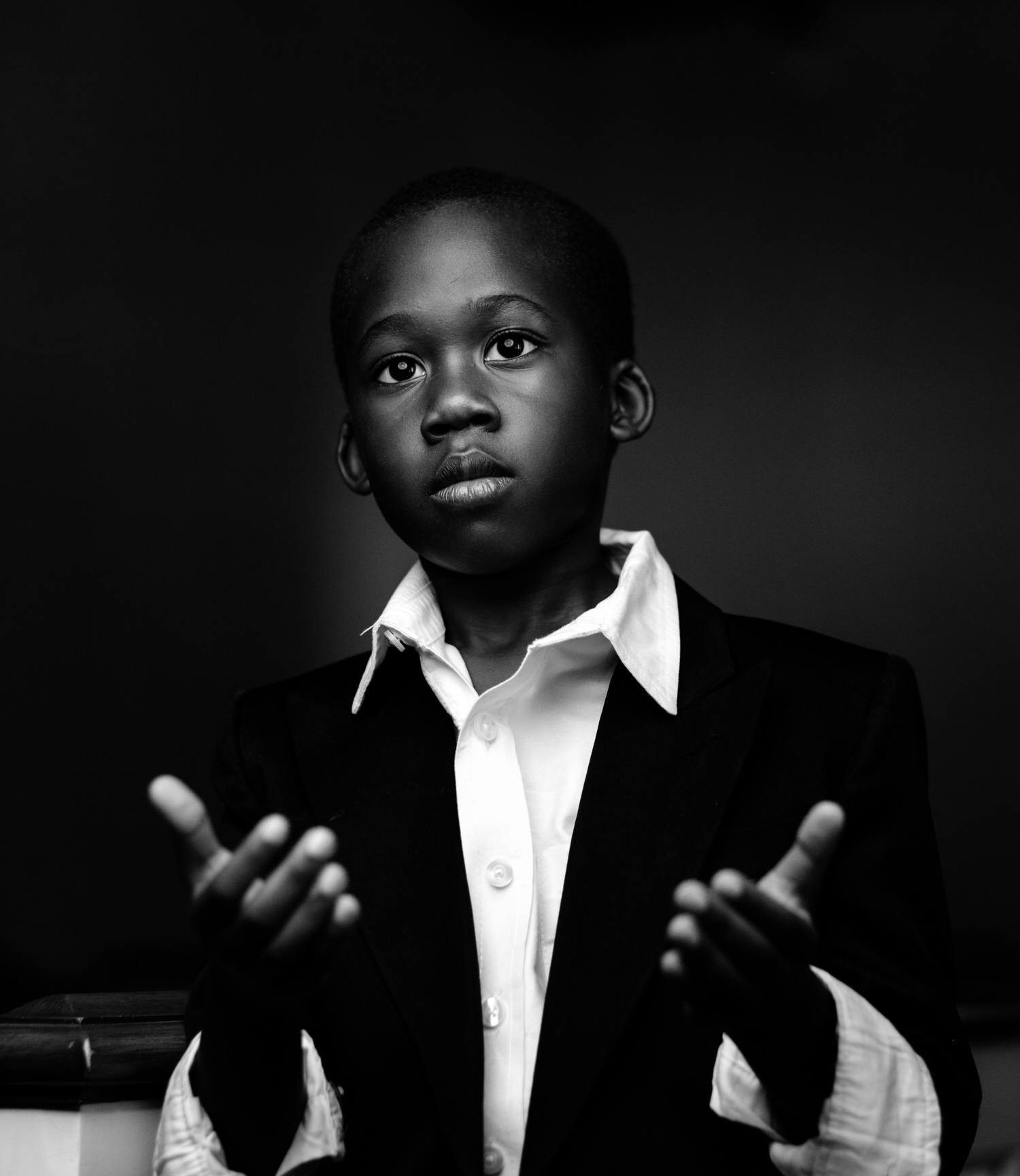 A boy in a blazer and open-collared shirt, hands raised, against a dark backdrop